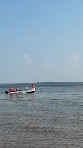 This video captures a serene day on the water in Krasnyi Bor, Russia. The primary focus is a small, white motorboat with a blue stripe, navigating a calm, dark body of water. Three individuals are visible in the boat, all wearing bright orange life vests, indicating safety precautions are being taken. The person in the front is sitting on the edge of the boat, facing away from the camera, while the other two are seated closer to the middle and rear of the vessel.

The boat is creating a gentle wake, suggesting it's moving at a moderate speed. In the distance, the horizon is marked by a hazy line of trees or land, under a vast, pale blue sky with a few scattered clouds. The overall atmosphere appears peaceful and relaxed, likely a leisure outing on a clear day. The time of day seems to be afternoon, given the bright, even lighting.

Visible text on the boat reads "P170-03УД". The water itself is rippled, reflecting the sky and creating a slightly textured surface. The scene evokes a sense of tranquility and simple enjoyment of nature and water activities.
