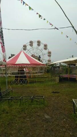 The video shows a dormant amusement park on a cloudy day. In the foreground is a small metal staircase leading to a swing carousel. In the background, there’s a Ferris wheel, a red-and-white striped tent, and another shelter-like structure. The ground is muddy and covered with grass. Colorful flags are strung above the scene. The park appears deserted and not in operation.