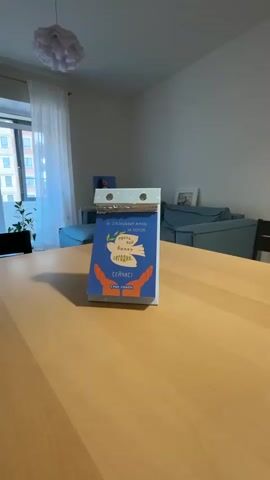 The camera shows a view of the corner of a dining room, focused on a table with a calendar in the center. The calendar has a blue cover with white and orange images and what appears to be a message in Russian. In the background, a sofa can be seen, as well as a window with white curtains. The room is lit with natural light. There is music playing, but no people are visible in the scene.
