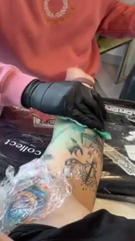 A person wearing a pink sweatshirt and black gloves is shown in the act of cleaning a tattoo on someone's arm with a green cloth. The arm has several tattoos and is covered in plastic wrap. There is a box under the arm with text on it. The person's arm and part of their torso are also visible.