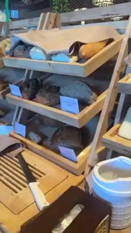 The video shows a wide selection of breads and pastries displayed on wooden shelves at a buffet. The breads include different types of sourdough, rolls, and loaves. The pastries include croissants, danishes, and muffins. There are also small bowls of chocolate squares. Each item has a label in front of it. A bread knife rests on a cutting board in front of the shelves. The shelves are arranged in a multi-tiered display.