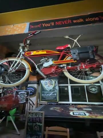 This video pans across the interior of what appears to be a motorcycle-themed bar or establishment in Kalai, Thailand. The scene is filled with various objects, primarily related to motorcycling culture.

Dominating the upper portion of the frame is a vintage red Indian motorcycle suspended from the ceiling, a prominent display piece. Below it, the walls are adorned with numerous motorcycle club logos, patches, and posters, many featuring fiery graphics and text like "You'll NEVER walk alone." There's a large, carved wooden elephant statue, also a significant focal point. Signs with phrases like "WELCOME OPEN!" and "RIDE HARD OR STAY" are visible, along with what looks like a menu or informational board with Thai script. Other decorative items include a fan, a cowboy hat, and various trinkets.

The atmosphere seems to be casual and edgy, typical of a biker bar. The lighting suggests it could be evening or nighttime, enhancing the moody ambiance. The presence of Thai script on some signs confirms the location as Thailand. No people are clearly visible or actively engaged in the scene. The overall impression is that of a space celebrating the freedom and camaraderie associated with motorcycle riding.