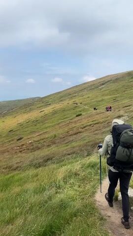 A hiking trail winds up a grassy mountainside under a partly cloudy sky. Several hikers are visible on the trail, some in small groups and others individually. The foreground is dominated by tall, green grass. A person wearing a backpack and using trekking poles is in the right foreground, facing away from the camera. The landscape appears expansive and natural.