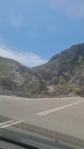 The video shows a car driving on a two-lane road through a mountainous area. The mountains are rocky and covered with some sparse vegetation. The sky is blue with a few clouds. Two cars are visible on the road ahead of the vehicle recording. The road has a solid double yellow line down the center. There are guardrails along the side of the road in some sections.