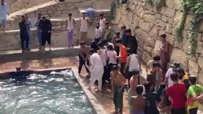 The video shows a group of approximately 30 young men and boys gathered around a small pool or pond surrounded by stone walls. Some individuals are in the water, while others are standing on the edge, and some are throwing each other into the water. The people are dressed in a variety of casual clothing. The scene appears to be taking place outdoors in a somewhat rustic or rural location. The lighting suggests it is a sunny day. The individuals seem to be engaging in playful behavior.