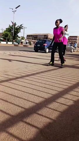 Here's a concise description of the video:

The video shows two young women roller skating on a paved area, likely a sidewalk or plaza, in an urban environment during daylight. The main focus is on the woman in the foreground, wearing a pink shirt and black pants, who attempts to skate. She is unsteady and uses her arms for balance. Behind her, another woman in a blue and pink top and black pants skates with more confidence. The setting includes a street with passing vehicles, buildings, trees, and other pedestrians visible in the background. A metal railing casts shadows across the pavement in the foreground.