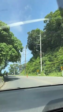 The video, shot from inside a vehicle, shows a two-lane road winding through a lush, green, and hilly environment. The road is asphalt with yellow lane markings. Trees with dense foliage line both sides of the road, and on the right, the terrain slopes upwards, covered in vegetation and dotted with electrical poles and wires. In the distance, several motorcycles are visible on the road. Traffic cones are placed on the road, and yellow warning signs indicating curves are present on the left side. The sky is blue with scattered clouds, and the sunlight suggests it is daytime. The vehicle's dashboard is partially visible in the foreground.