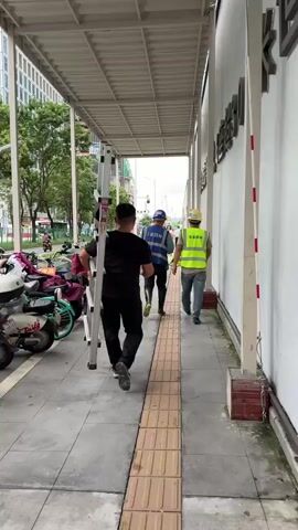 The video shows a pedestrian walkway with several people. In the foreground, a man in a black shirt and black pants walks away from the camera, carrying a silver ladder horizontally over his right shoulder. He is walking on a sidewalk with tactile paving. Behind him, two other men in construction vests and hard hats are also walking away from the camera. The man in the middle wears a blue vest, and the man on the right wears a bright yellow-green vest. To the left of the sidewalk is a row of parked scooters. Further in the background, two women are seen, one on a bicycle and another walking. The environment appears to be an urban area with buildings and trees visible. The walkway is covered by an overhead structure.