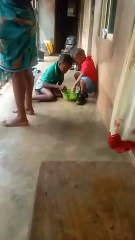 Two young boys are on the floor looking into a green plastic bowl. The boy on the left wears a green shirt and is kneeling, while the boy on the right wears a red shirt and is sitting, leaning against a wall. A woman in a blue and green printed skirt stands near them. A wooden bench is in the foreground, and a curtain hangs near the wall behind the boys.