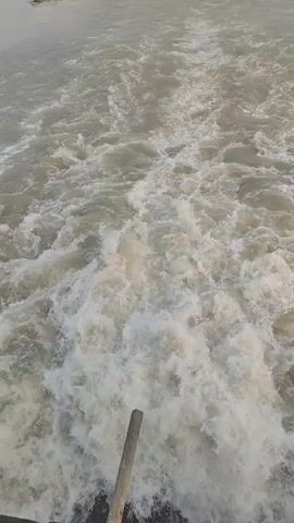 The video shows the wake of a boat, creating foamy waves in the water. A smaller boat with people on board is visible in the distance, ahead of the camera. The water appears grey and choppy. A portion of a wooden pole is visible at the bottom of the frame, likely part of the vessel from which the video is being taken. The sky is overcast and hazy.