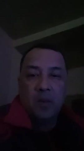 A medium-shot video shows a man with short dark hair. He's wearing a dark jacket with a red collar. He is indoors, likely at home. The room is dimly lit with green accents. He is looking at the camera and speaking. He initially holds up his hand, then lowers it. He appears to be addressing someone off-camera or recording a message.