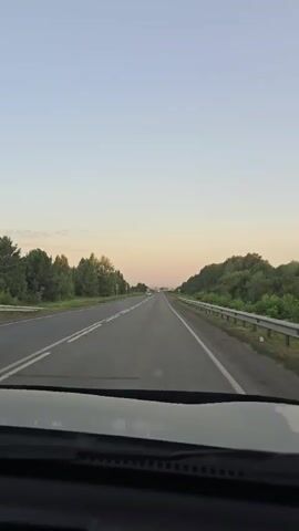A first-person perspective video shows a car driving on a paved road at dusk. The road has two lanes in each direction, separated by a white line. Trees and greenery line both sides of the road. A guardrail is visible on the right side of the road. In the distance, the sky is a pale orange and pink hue, indicating the setting sun. There are a few other vehicles on the road, including a truck in the distance on the left lane and cars in the right lane in the distance. Some signs are visible on the side of the road. The interior of the car, specifically the dashboard, is visible in the foreground.