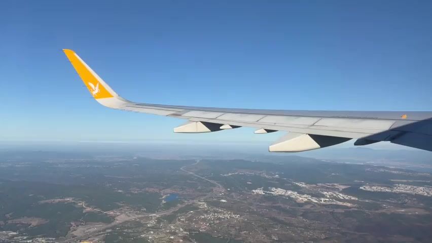 A wing of a Pegasus Airlines airplane is visible extending to the left of the frame. The wing is gray with a yellow tip featuring the Pegasus logo. The airplane is flying over a landscape under a clear blue sky. The ground below is a mix of green vegetation, scattered buildings, and a road. The horizon is visible in the distance, with a hazy line where the sky meets the land.