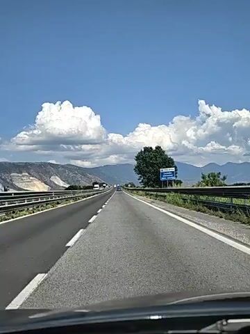 The video shows a first-person perspective of driving on a highway. The road is asphalt with white lane markings. On the left side of the road, there is a metal guardrail and a sloping embankment with some vegetation. On the right side, there is also a guardrail and more vegetation. In the distance, mountains are visible under a bright blue sky with scattered white clouds. Several vehicles are on the highway, including cars and trucks. There is a single large tree on the right side of the road, and a blue sign with white text is visible behind it. The camera appears to be mounted inside a vehicle, possibly on the dashboard, as the lower edge of the windshield and wipers are visible. The footage is shaky, indicating movement.
