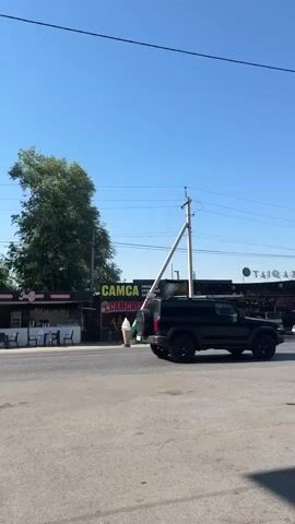 The video opens showing a tilted utility pole leaning over a street, with power lines running across the frame. A black SUV is parked on the right side of the street, facing away from the camera. Behind the SUV, several other cars are visible. On the left side of the street, there are small shops with signs that read "CAMCA". A tall, conical ice cream cone sculpture is placed in front of one of the shops. The sky is clear and blue, indicating a sunny day. The camera then pans to the right, revealing more shops and a large, covered outdoor area with lights strung overhead. People are seen walking in the background. The video ends with a shot of a white SUV parked on the right side of the street, facing towards the camera.