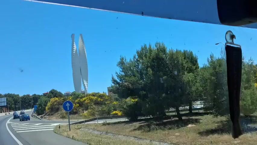 The video is taken from the interior of a vehicle, likely a truck or bus, as it drives along a highway on a sunny day. The sky is clear and blue. Initially, a large, white, abstract sculpture is visible on a hill to the left of the road, along with a building in the background and yellow flowering bushes. The view then shifts, showing the highway with multiple lanes of traffic. A blue semi-truck is driving in the same direction, and other cars are visible. There are also green trees and bushes alongside the road. The road appears to be well-maintained, and street lights are visible. Road signs are also present.