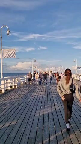 The video shows a wooden pier with many people walking along it. The pier extends into the water, with the ocean visible on the left side. The sky is blue with scattered clouds, and the sun is shining, casting long shadows on the wooden planks. Several people are walking towards the camera, while others are walking away. Some are in pairs, while others are in small groups. The pier is lined with white railings and benches, and lampposts are spaced along its length. The overall atmosphere appears to be relaxed and pleasant, with people enjoying a stroll along the waterfront.
