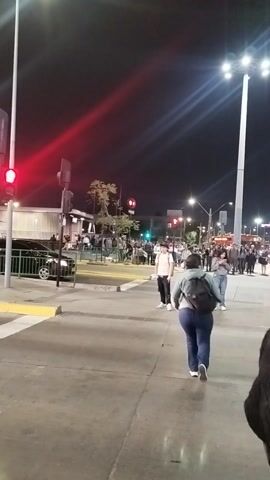The video captures an urban street scene at night in Maipú, Chile. The perspective is from a stationary position, likely on a sidewalk or curb, looking across a street.

In the foreground, a man wearing a red jacket with a white Adidas logo, a black beanie, and dark pants walks from left to right, partially obscured by a passing silver/white car. Another dark-colored car is briefly visible before him.

The background shows a busy street with multiple people gathered on sidewalks. A line of streetlights illuminates the area, casting bright beams and flares. Traffic lights are visible, with one showing red and another green, indicating an intersection. Buildings and other structures can be seen further in the background. The overall atmosphere suggests a typical evening in a city, with pedestrians and vehicles moving about. No specific event or clear emotions are discernible from the footage.