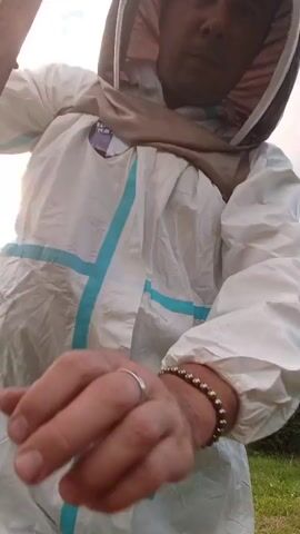 A beekeeper in a white protective suit with blue accents holds up a wooden frame filled with bees and honeycomb. The beekeeper is a person with short hair and is looking at the camera. The background shows a grassy area with some bushes and trees, and the sky is overcast. The video is shot in a close-up, focusing on the beekeeper and the frame of bees.