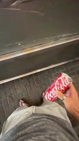 A first-person perspective video shows a person holding a can of Coca-Cola. The person is wearing flip-flops and light-colored shorts. They are standing on a tiled surface in front of a dark wall. The video then transitions to a street scene at night. A blue car is parked on the side of the road. There are people standing on the sidewalk, some are talking to each other. String lights are visible above the sidewalk, illuminating the scene. The person is now walking in the street, and the camera is moving with them, showing the environment from a low angle. The overall scene suggests a casual, outdoor evening setting, possibly in a commercial area.