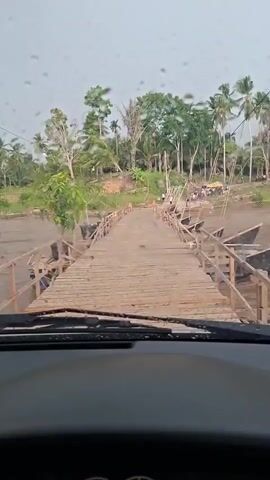 The video shows a view from inside a vehicle looking forward at a wooden bridge extending over a body of water. The bridge is made of planks and has railings on either side. There are several boats docked on the right side of the bridge. On the far bank, there are trees and some people gathered, possibly at a market or gathering. The weather appears overcast, and there are raindrops visible on the windshield. The vehicle appears to be moving forward onto the bridge.