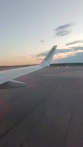 A passenger on a Ryanair airplane films a wide, static shot from their window seat, looking out at the aircraft wing, the airport tarmac, and the horizon. The airplane wing, white with "RYANAIR" written in blue on the wingtip, occupies the left side of the frame. The tarmac, dark grey, stretches across the foreground and midground, with yellow lines visible on the surface. In the distance, bright lights are visible, indicating an airport runway or buildings. The sky above is a gradient of light blue, pink, and orange, suggesting either sunrise or sunset. A few scattered clouds are present. The only sound is a female voice speaking in a foreign language.