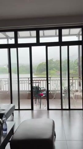 The video begins with a view of a rainy scene outside a sliding glass door. The camera then pans to the right, revealing a living room interior. A Buddha statue wearing sunglasses and a hat sits on the floor. A white angel statue is on a small ornate table. A television is on a low gray console. The flooring is light-colored tile. The wall is white.