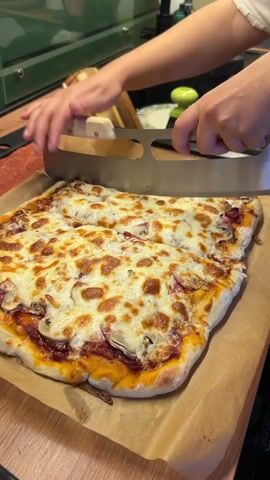 In a kitchen setting, likely in Shepelyovo, Russia, a person is actively engaged in cutting a rectangular, golden-brown pizza. The pizza is generously topped with melted mozzarella cheese, mushrooms, and red onions. The cutting is being performed with a specialized pizza cutter featuring a large, curved blade and a wooden handle. The pizza rests on a sheet of parchment paper, which is placed on a wooden countertop. The background reveals a kitchen sink, a wooden cutting board, and various kitchen utensils, including a bottle of pink liquid and a green-handled grater. The lighting suggests it could be daytime. The overall scene conveys a domestic and culinary activity. There is no visible text in the media.