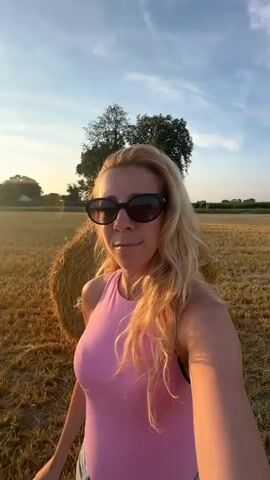 A blonde woman wearing sunglasses and a pink top is taking a selfie video in a field of hay bales at sunset. She is smiling and gesturing with her right arm towards the setting sun. The field is dry and golden, with rows of cut straw. Several large, round hay bales are visible in the background, as well as trees and a clear blue sky. The sun is low on the horizon, casting a warm, orange glow over the scene.
