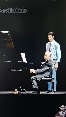 A man in a suit sits at a piano with a young man standing behind him. The seated man is looking at sheet music on the piano. The standing man places his hands on the seated man's shoulders. The seated man turns his head to look at the standing man. The seated man then gestures to the sheet music with his left hand. The standing man then moves his hands down.