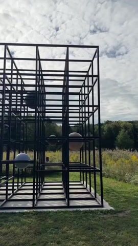 The video shows a slow pan of a large, black metal cube sculpture outdoors. The sculpture is made of interconnected square frames, creating a grid-like structure. Inside the structure, several spheres of varying sizes and colors are suspended. The background consists of a grassy field with tall, dry grass and trees in the distance. The sky is cloudy. The camera movement is smooth and steady, providing a comprehensive view of the sculpture and its surroundings. There are no people visible in the video, and no interactions are captured. The lighting suggests it is daytime, with diffused light from the overcast sky.
