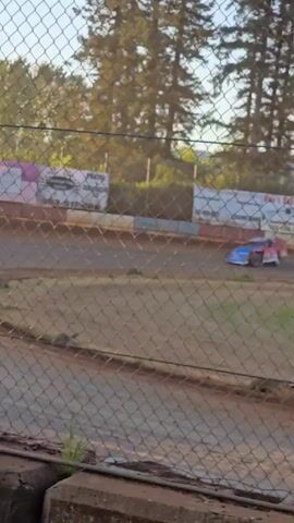 Here is a description of the video:

The video shows a dirt track race with several modified race cars speeding around a turn. The cars are mostly blue and white, and they kick up a lot of dust. The video is taken from behind a chain-link fence, with trees and advertising signs visible in the background. The weather appears to be sunny.
