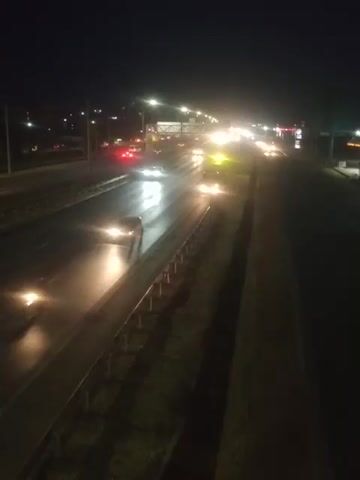 The video shows a nighttime view of a multi-lane highway with heavy traffic. The camera angle is high, looking down at the road from an elevated position, possibly a bridge or overpass. The headlights and taillights of numerous vehicles create streaks of light on the wet asphalt, indicating that it may be raining or has recently rained. The traffic appears to be moving slowly, suggesting a traffic jam or congestion. The surrounding environment is dark, with only the artificial lights of the highway and distant city lights visible. There are also lights along the side of the highway, likely streetlights. The overall scene depicts a common urban nighttime driving experience characterized by illuminated traffic on a dark roadway.