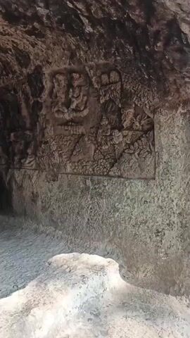 The video shows the interior of a rock-cut cave, likely a temple or historical site. The cave walls are rough and uneven, with some sections appearing to be carved or sculpted. In several frames, intricate carvings of figures, possibly deities or historical figures, are visible on the walls. The floor of the cave is covered with small, light-colored stones or gravel. The lighting is dim, suggesting natural light filtering in from an unseen entrance or artificial lighting that is not prominent. The camera movement is slow and panning, allowing the viewer to observe the details of the carvings and the cave structure. There are no people visible in the video, and no clear objects other than the natural rock formations and the carvings themselves. The overall impression is one of ancient architecture and religious or historical significance.