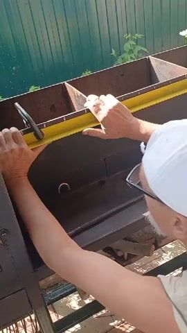 A person wearing a white baseball cap and glasses is working on a metal structure. They are holding a yellow measuring tape and using a tool to adjust something within the structure. The person's hands are visible, and they appear to be focused on the task. The background shows a green fence and some greenery. The overall scene suggests an outdoor work environment.