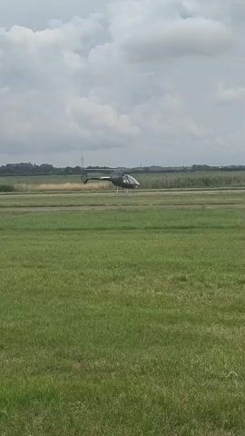 A dark helicopter is on a runway, its rotors spinning. The helicopter appears to be stationary or moving very slowly. The runway is bordered by green grass. In the background, there are fields, trees, and a cloudy sky with a few windmills visible. The lighting suggests it is daytime.