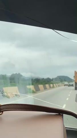 The video is taken from inside a vehicle, looking out through the windshield. The windshield is cracked. In the foreground, the top of a brown purse is visible. The vehicle is traveling on a highway. There are other vehicles on the road in the distance. The road is lined with a barrier of alternating yellow and black striped concrete blocks. Beyond the barrier, there is greenery and hills or mountains in the distance. The sky is overcast with clouds. The overall impression is that of traveling on a highway under cloudy skies.