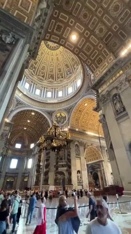 A low-angle shot showcases the grand interior of a church, filled with tourists. The camera slowly pans right, revealing the vast, opulent space adorned with gilded ceilings, intricate frescoes, and massive statues. Sunlight streams through the high windows, illuminating the details of the architecture and the ornate altar, while tourists mill about, admiring the surroundings.