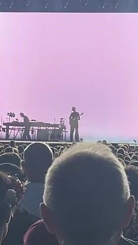 A concert performance is underway in a large venue, observed from the perspective of an audience member. On stage, three individuals are visible, positioned behind a long, illuminated console. The person on the left plays a guitar, while the person in the center appears to be operating equipment on the console. The person on the right also holds a guitar. The stage is lit by a bright, white backdrop that shifts to a soft pink at 00:01 and then transitions to a dark, smoky atmosphere with spotlights at 00:02. The foreground is filled with the heads and shoulders of the concert-goer's immediate surroundings.