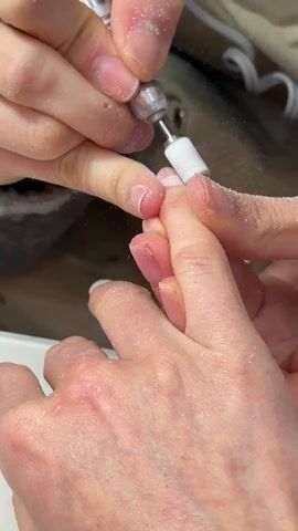 Here is a description of the video. 
The video shows a person getting a manicure. The manicurist is using a small electric drill to smooth the surface of the customer's fingernail. Small particles of nail are flying into the air. The environment appears to be inside a salon.