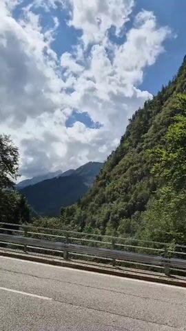 This is a POV video from a vehicle driving along a winding road through a mountainous, forested area. The road is paved, and there is a guardrail on the left side of the road. The mountains are steep and covered in dense green trees. The sky is blue with fluffy white clouds. As the video progresses, the road curves more sharply, and the view opens up to reveal more of the dramatic mountain landscape and the valley below. In the last few seconds, another car, a small orange one, is seen driving around the bend ahead.
