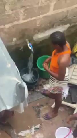 Two women are filling buckets with water from a faucet in an outdoor area with a concrete floor. One woman is wearing an orange tank top and floral shorts. The other woman is wearing a white tank top and a blue crocheted head covering. A large gray sheet partially covers one side of the scene. There are several buckets, including black, green, and white. The area appears to be an outdoor utility space, possibly for water collection or cleaning.  The brick wall behind the faucet has some dampness or moss.