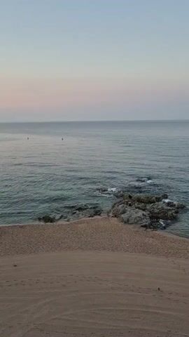The video shows a serene beach scene at dusk. The sky transitions from a pale blue to a soft pink near the horizon. The sea is calm with gentle waves lapping at the shore. In the middle distance, a few scattered rocks emerge from the water, creating a small cove. The beach itself is sandy and appears mostly empty, with faint footprints visible in the sand. The overall atmosphere is peaceful and tranquil.