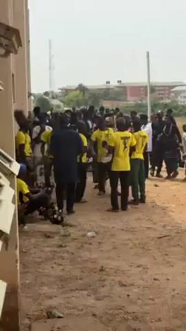 The video shows a group of people gathered outside a building. Many of them are wearing yellow t-shirts with a black graphic on the back. Some individuals are playing drums, and others are dancing. There is a man in a dark suit who appears to be directing the group. The ground is dirt and there is a building in the background.
