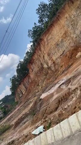 The video shows a large, steep dirt embankment. The top of the embankment is lined with trees, and the bottom is covered with loose dirt and vegetation. There are power lines in the foreground and a few buildings at the base of the embankment. The sky is blue with some white clouds. The camera is angled upwards, giving a perspective of the height and scale of the embankment.
