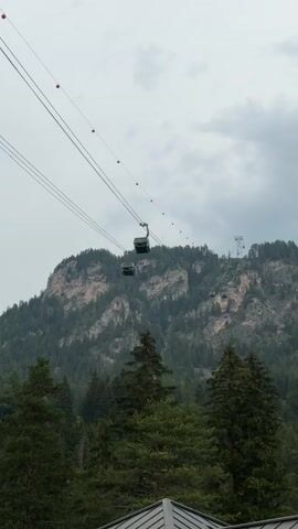 The video shows a cable car system ascending a mountainside. In the foreground, the roof of a building is visible. Behind the building, a dense forest of evergreen trees covers the lower slopes of the mountain. The upper part of the mountain is rocky and appears to be barren. Several cable cars are seen moving up the mountain along suspended cables. Red markers are attached to the cables at intervals. The sky is overcast with clouds. The overall impression is of a scenic mountain landscape with a functional cable car infrastructure.