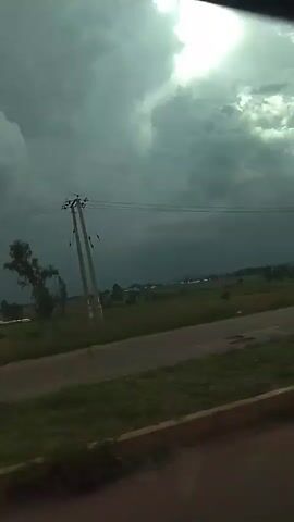 The video pans across a cloudy sky with the sun partially visible through the clouds, creating a bright spot. Below the sky, there is a landscape with green fields, some trees, and a road. Power lines and a utility pole are visible. The overall impression is of a somewhat overcast or stormy day.