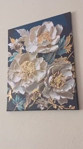 The video shows two framed paintings hanging on a beige wall. The first painting, on the left, depicts three large white and cream flowers with golden accents on a dark blue background. The second painting, on the right, displays a cluster of round, fluffy flowers in various vibrant colors like pink, orange, yellow, and blue, against a dark, leafy background. The camera slowly pans from the first painting to the second.