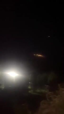 The video shows a nighttime scene with a bright light on the left side of the frame, possibly from a vehicle or streetlamp. In the distance, there is a cluster of lights, appearing as a faint orange glow. The environment seems to be outdoors with trees or bushes visible in the foreground and midground, silhouetted against the dark sky. There are no people visible in the video. The overall impression is of a rural or semi-rural setting at night.