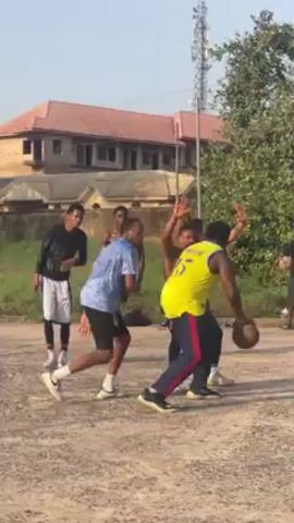 A group of five men are playing basketball on an outdoor court. One man, wearing a yellow jersey with the number 15, drives towards the basket and jumps to take a shot. Another man, wearing a blue shirt, attempts to block him. The shot is unsuccessful. Two other men stand nearby, watching the play. The basketball court appears to be unpaved, with a metal basketball hoop and backboard. In the background, there are trees, a building with a red roof, and a utility pole. The sky is clear and sunny.